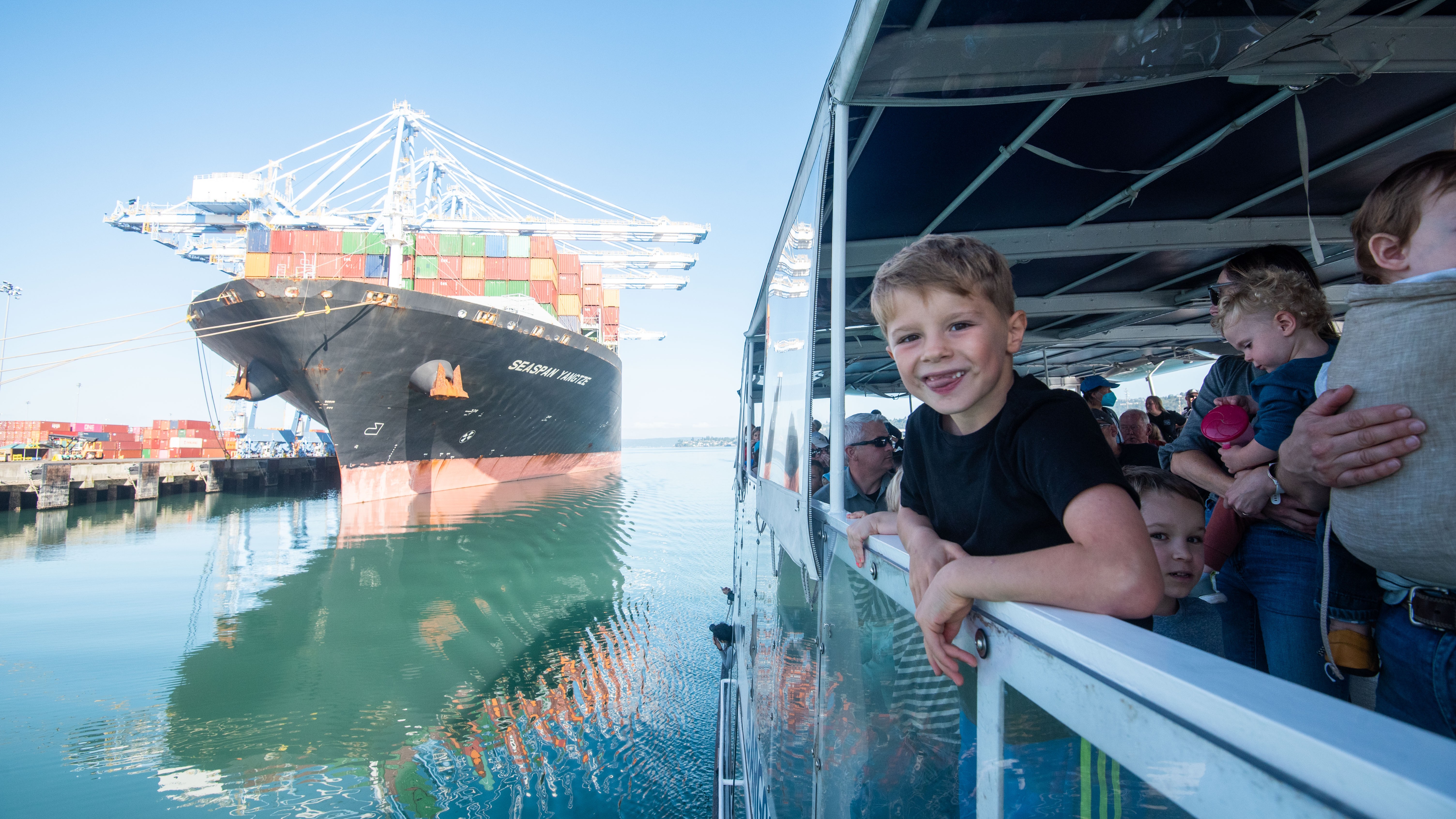 A young boy leans over the railing of a boat while grinning.