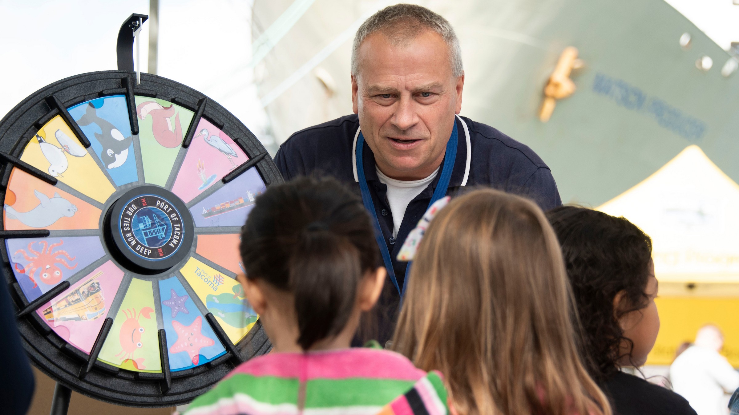 A man leans over to speak to children in front of a prize wheel at a Port event.