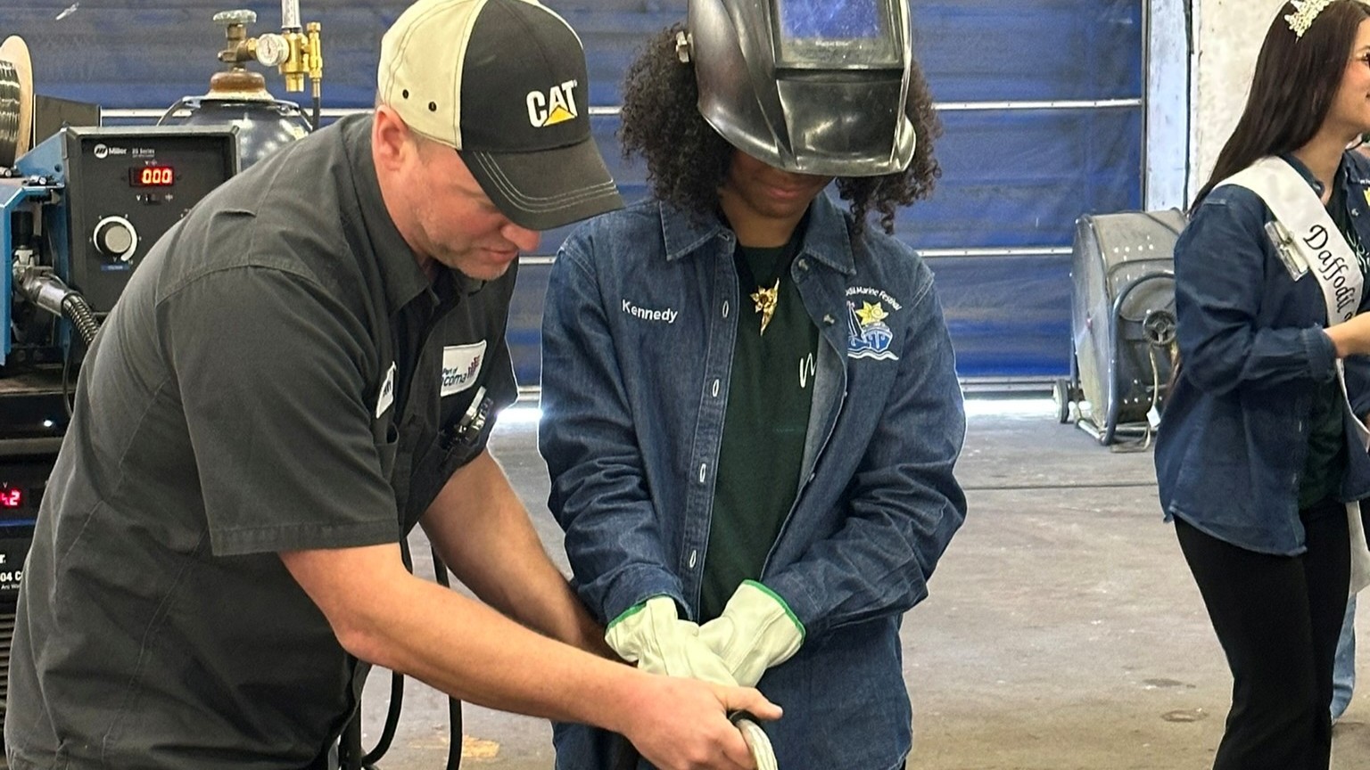 An adult male employee helps a student in protective gear with welding.